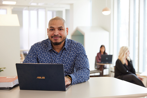 A man with his laptop on a desk with other people in the background