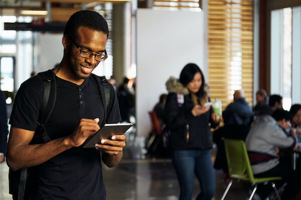 A man wearing a backpack looks at his cellphone. A group of students at table in are seen behind him.