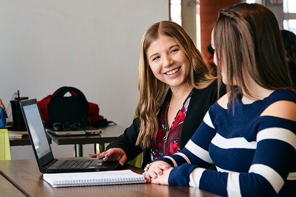 Two students talk while sitting at a table.