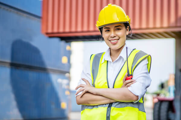A woman wearing a hard hat and safety vest crosses her arms and smiles at the camera.