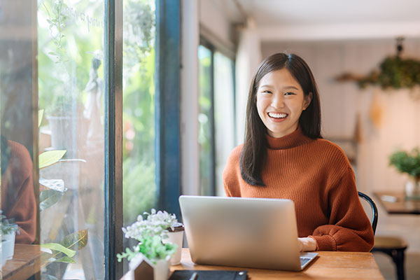 A woman smiles at the camera while working on her laptop in her home office.
