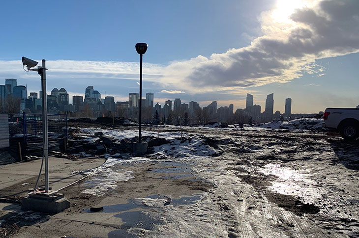 View of Campus Centre construction site from the SAIT Parkade entrance showing snow and ice on the flat dirt site with a lampost in the foreground and the Calgary downtown skyline in the background.