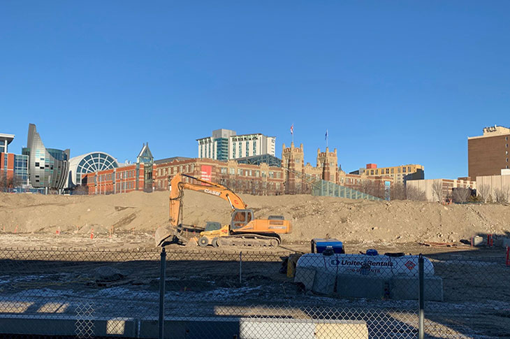 View of Campus Centre construction site from the CTrain platform. Large machinery on site moving earth. Other campus buildings are partially visible in the background, with mostly dirt mounds in the foreground.