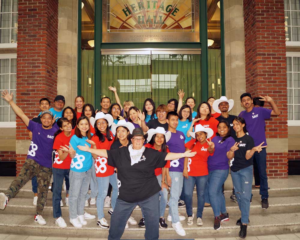 Hotel and Tourism Management students wearing white cowboy hats pose on the steps to Heritage Hall in the Irene Lewis Atrium, July 2022