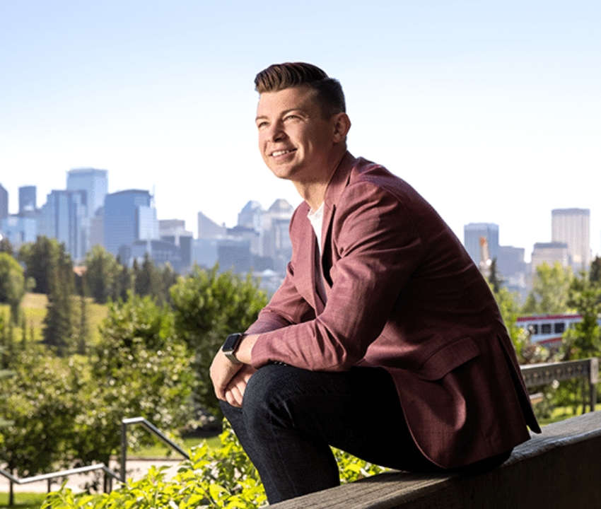 Kyle Bukauskas sitting on concrete wall ledge with city skyline behind him