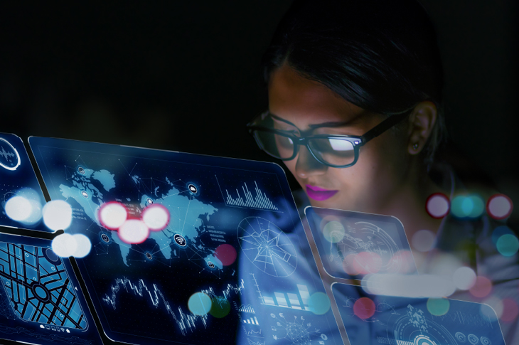 A woman wearing glasses looks at transparent screens with a world map graphic.